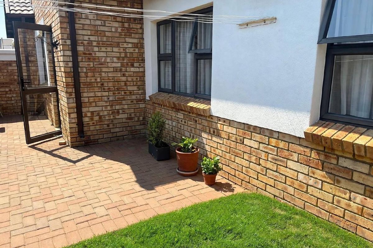 A well-maintained rear exterior area featuring a paved patio, brick and plaster facade, and a small lawn with aluminium-framed windows.