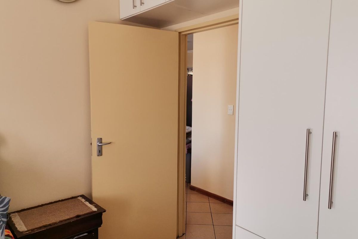 A bedroom interior featuring built-in white cupboards with overhead storage and tiled flooring, leading into an adjacent hallway.