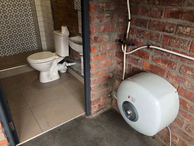 A functional bathroom featuring a toilet and basin, adjacent to an utility area with an exposed brick wall and electric water heater.