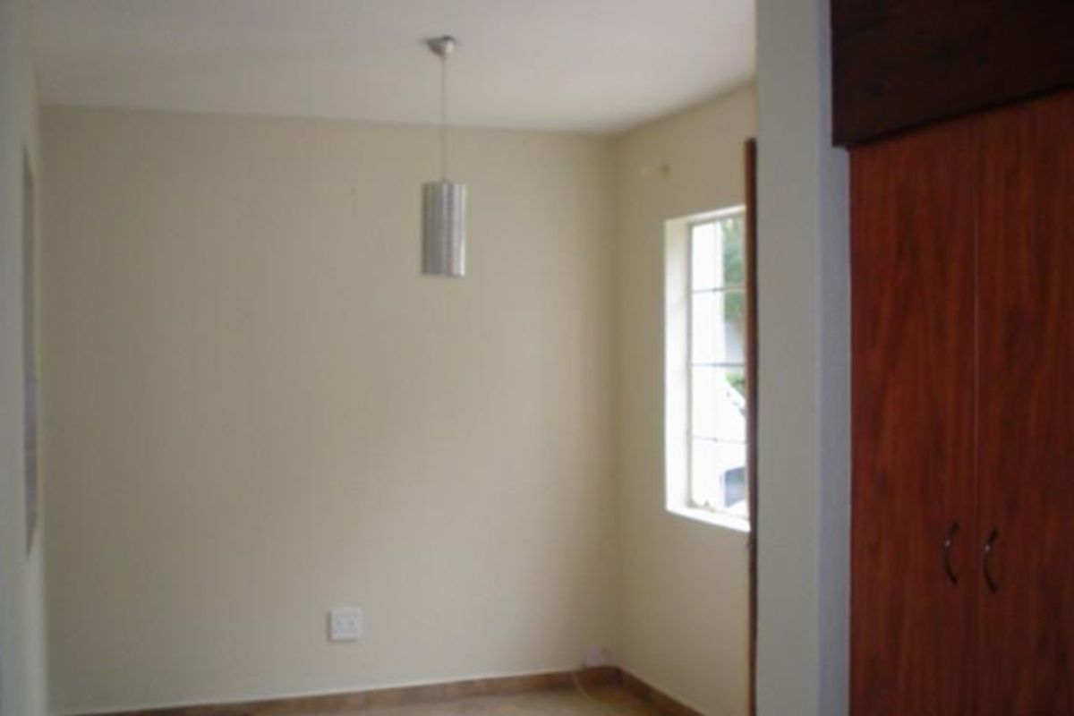 A simple, empty bedroom featuring tiled flooring, a built-in wooden wardrobe, and a window with security bars.