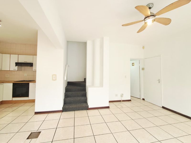 Open plan living area featuring tiled floors, a ceiling fan, and an adjoining kitchen with built-in cabinetry and appliances, leading to an internal staircase.