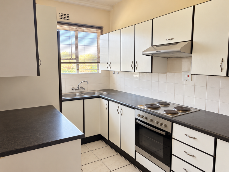 A functional kitchen featuring white cabinetry, dark laminate countertops, a built-in oven with hob, extractor fan, and tiled backsplash with natural light.