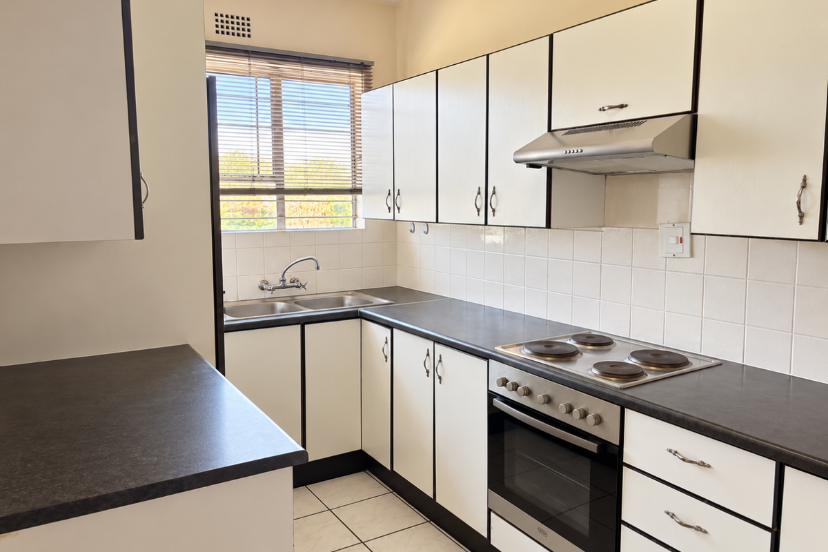 A functional kitchen featuring white cabinetry, dark laminate countertops, a built-in oven with hob, extractor fan, and tiled backsplash with natural light.