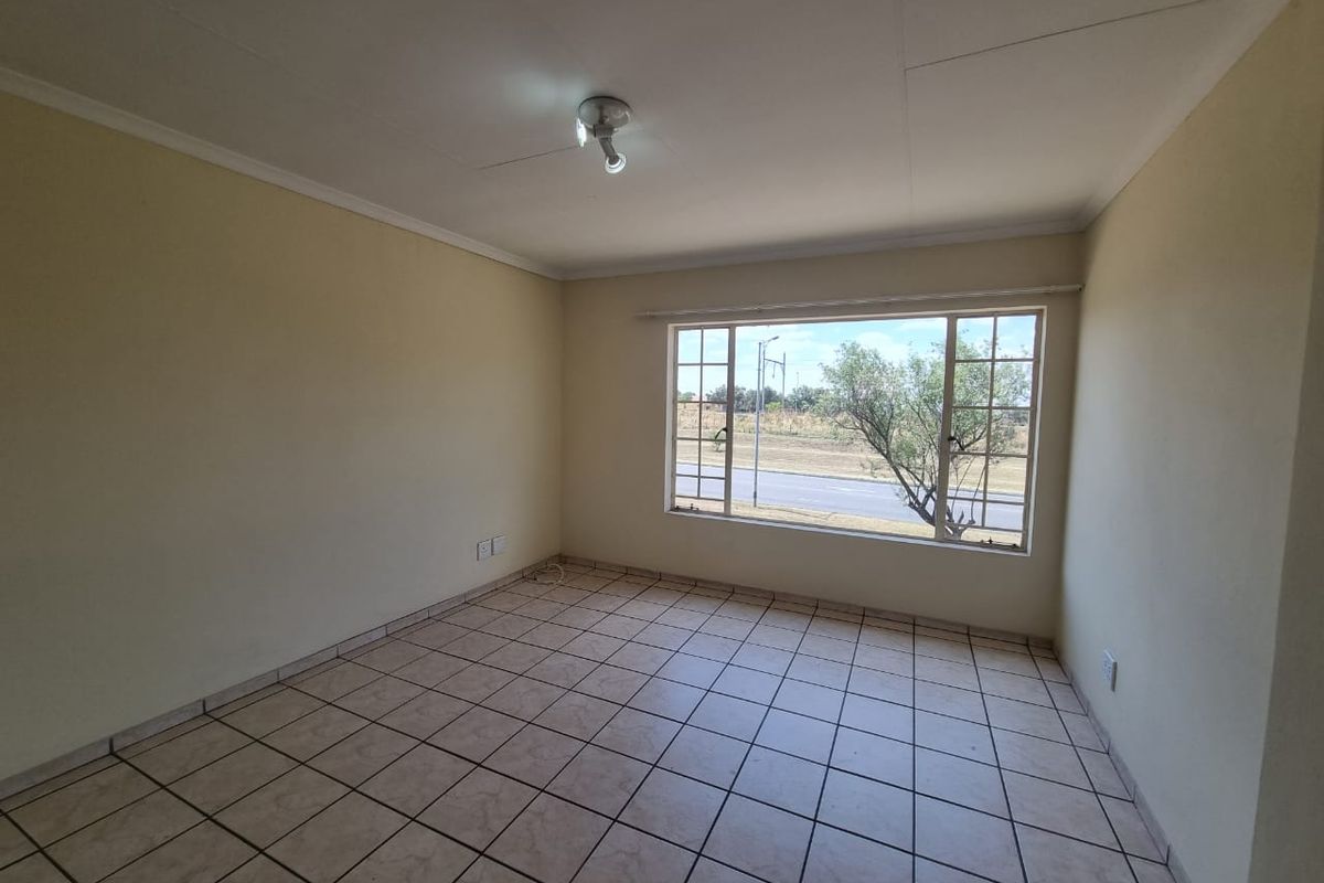 A bright, empty living room featuring neutral tiled flooring and a large window providing natural light and street views.