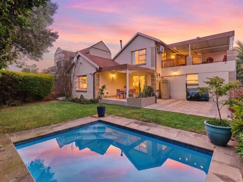 Rear view of the property featuring a swimming pool, paved patio area, covered veranda, and an upper-level balcony set within a landscaped garden.