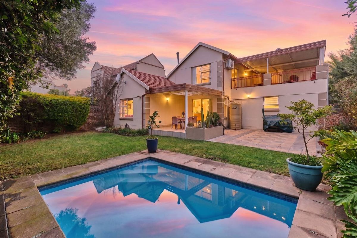 Rear view of the property featuring a swimming pool, paved patio area, covered veranda, and an upper-level balcony set within a landscaped garden.