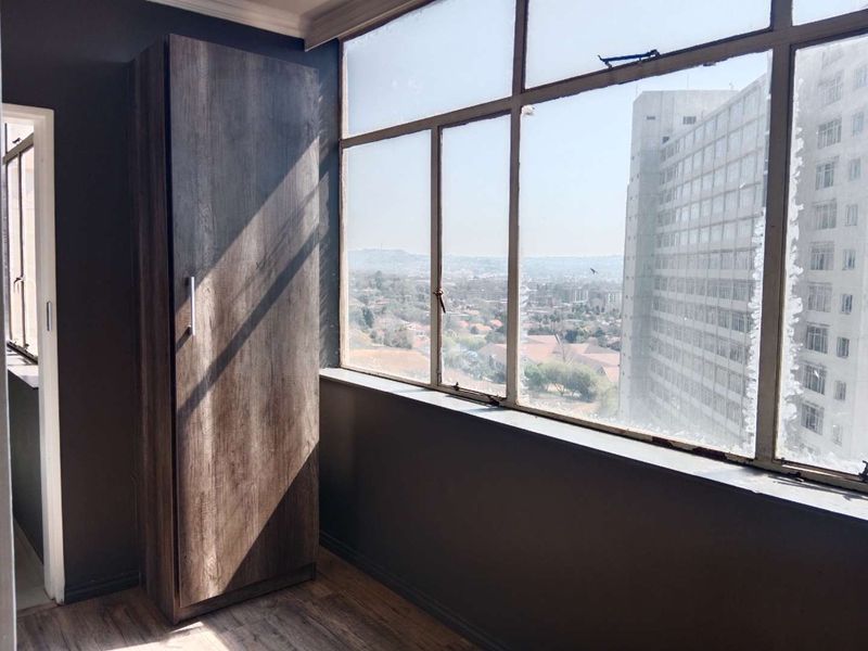 A bright bedroom featuring a built-in cupboard, laminate flooring, and large windows offering expansive city views.