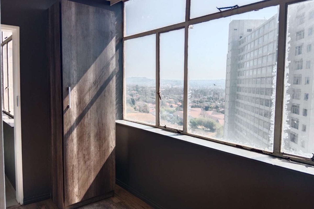 A bright bedroom featuring a built-in cupboard, laminate flooring, and large windows offering expansive city views.