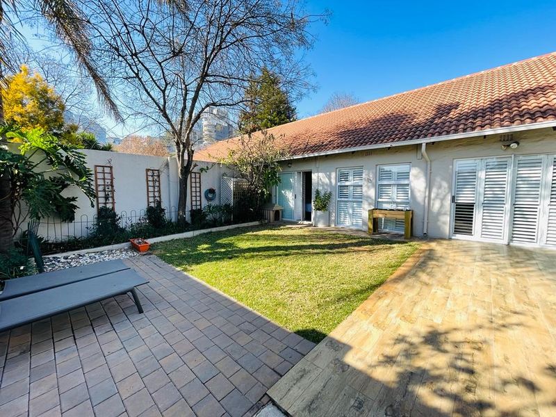 A well-maintained rear garden featuring a combination of paver and wood-look tiled patio areas, lawn, and secure white shutters on the building facade.