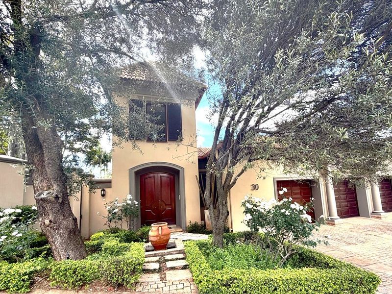 Front elevation of a Mediterranean-style residence featuring a double garage, paved driveway, and established landscaped garden with mature trees.
