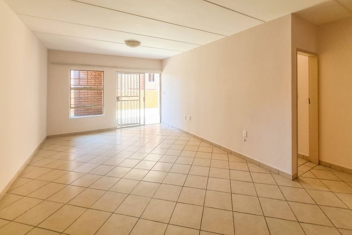 A bright, empty living room featuring neutral tiled flooring, a sliding glass door with security gate, and access to an adjacent area.