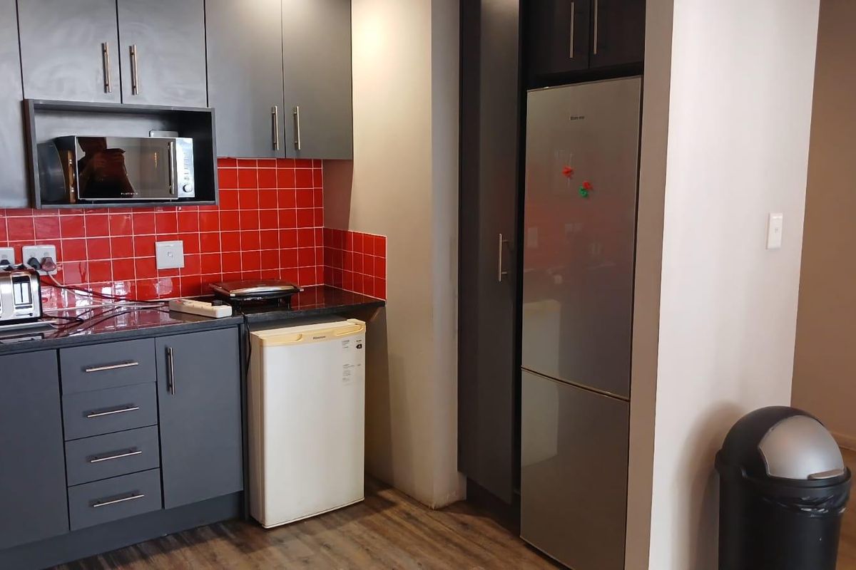 Compact kitchen featuring dark grey cabinetry, contrasting red tiled backsplash, granite countertops, and integrated storage space for a refrigerator.