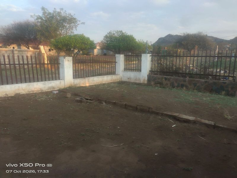 An outdoor garden area featuring perimeter metal fencing with concrete pillars and a stone retaining wall, currently requiring landscaping and ground maintenance.