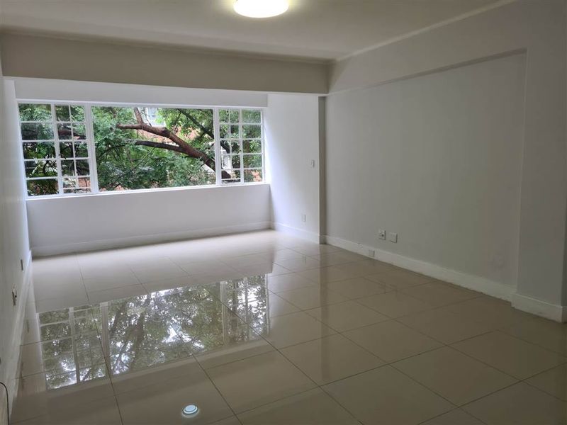 A bright, spacious, and empty living area featuring polished large-format floor tiles and a large multi-pane window with a leafy green outlook.