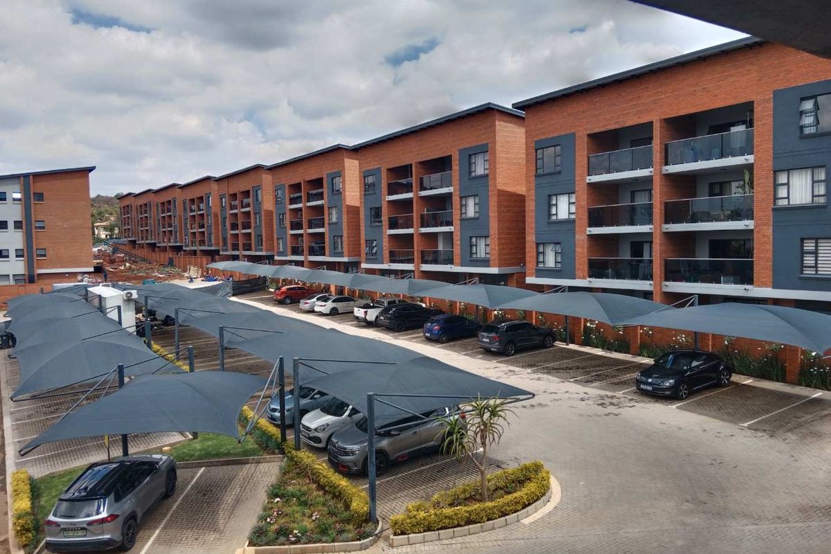 Exterior view of a modern apartment complex featuring brick facades, private balconies, and designated paved parking bays with shade cloth structures.