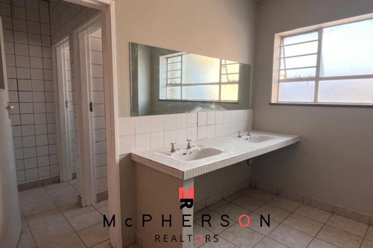 A functional office bathroom featuring a double vanity with tiled countertop, wall-mounted mirror, and tiled flooring, requiring cosmetic updates to modernize the space.