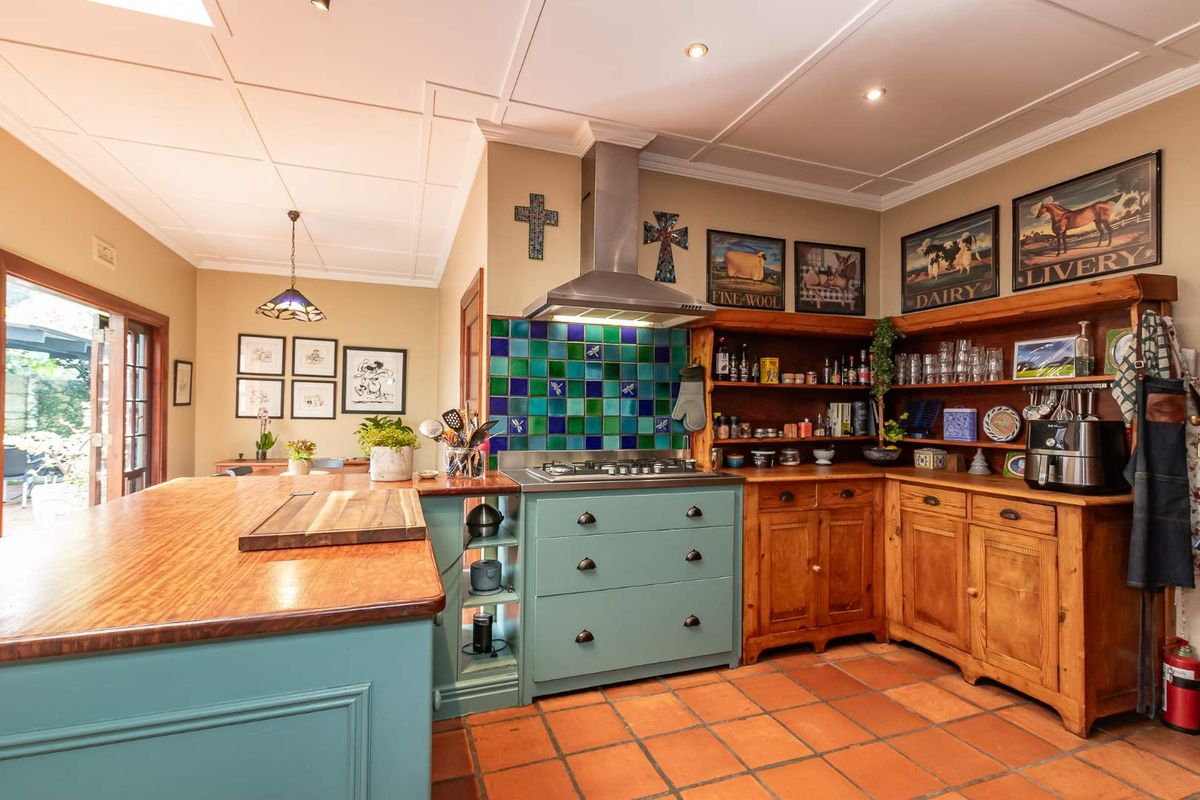 A rustic-style kitchen featuring a central island, gas hob with stainless steel extractor, vibrant tiled splashback, and warm terracotta flooring.