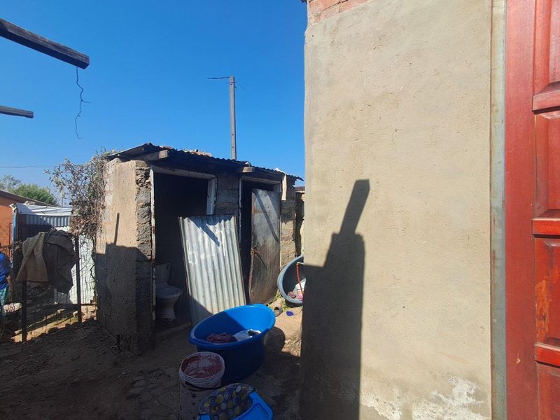 Rear exterior view showing a basic outdoor toilet structure with masonry walls and corrugated metal roofing in a state of significant disrepair.