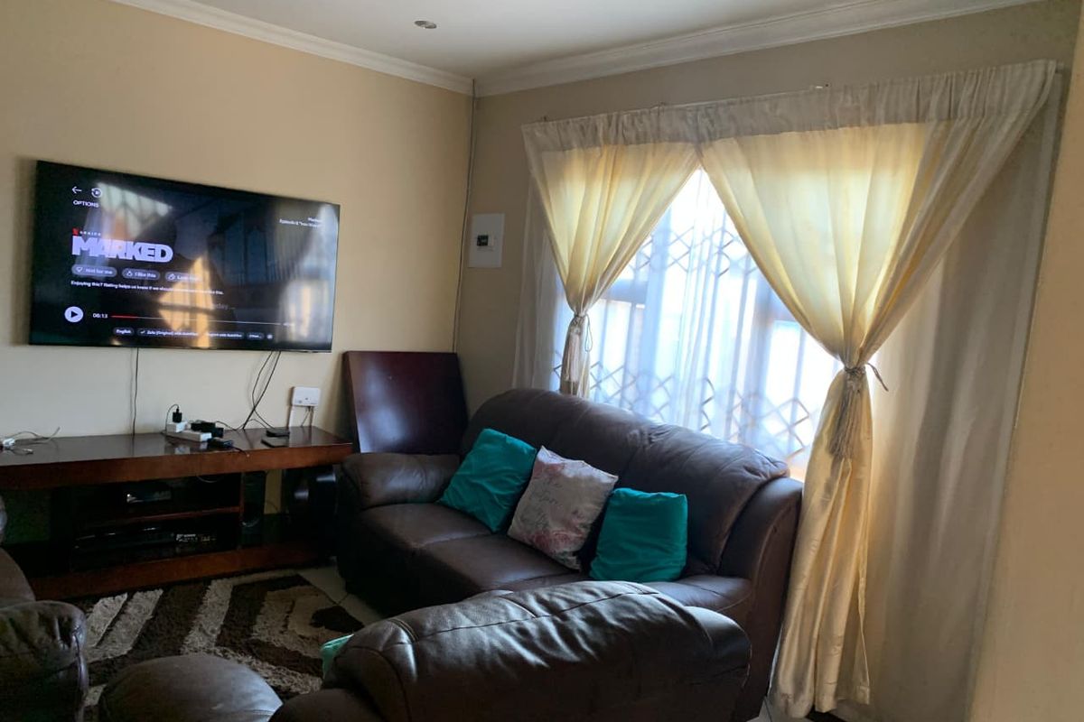 A cozy living room featuring tiled flooring, a wall-mounted television, and a window fitted with security bars and curtains.