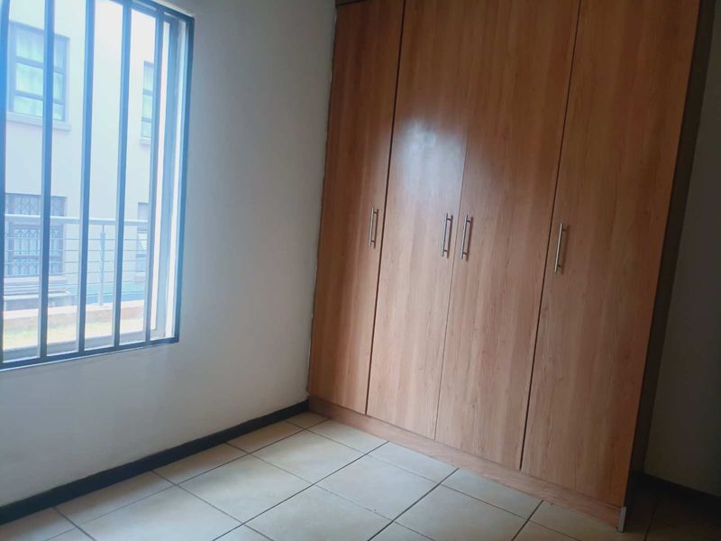 A clean, empty bedroom featuring light wood-finish built-in cupboards, neutral tiled flooring, and a window fitted with security bars.