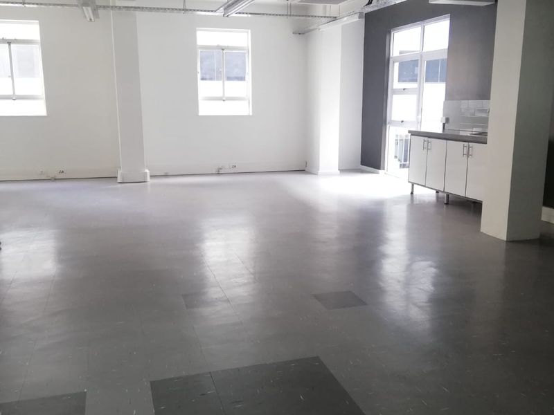 Spacious open-plan office area featuring exposed ceiling infrastructure, vinyl flooring, large windows for natural light, and a compact built-in kitchenette.