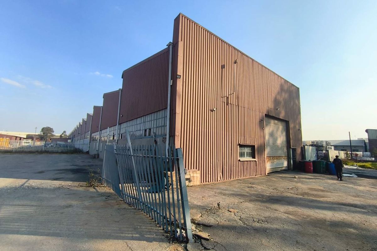 Industrial warehouse facility featuring corrugated metal cladding, a large roller shutter door, and an expansive asphalt yard area with perimeter security fencing.