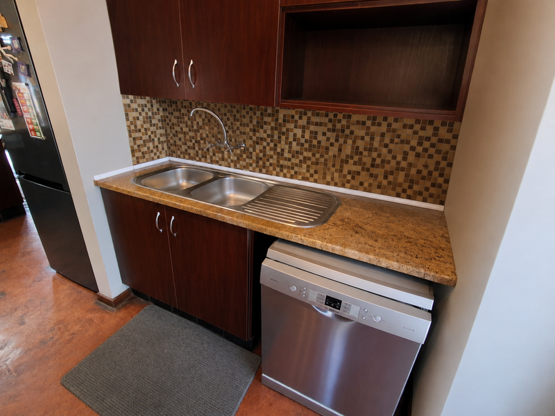 A functional scullery area featuring dark wood cabinetry, a double bowl stainless steel sink, mosaic tile splashback, and space for a dishwasher.