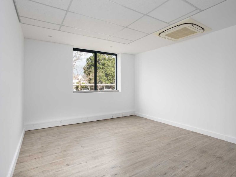 A bright, vacant office space featuring modern laminate flooring, a suspended ceiling with integrated air conditioning, and a large window providing natural light.