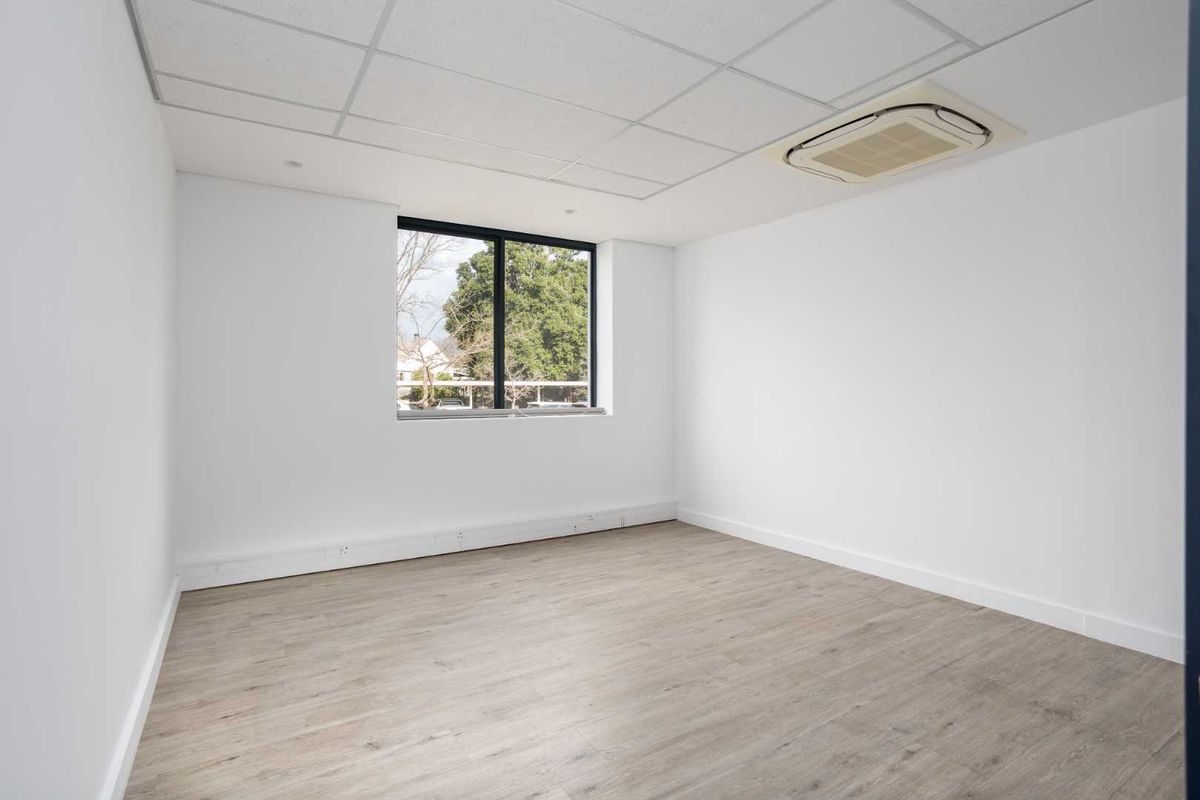 A bright, vacant office space featuring modern laminate flooring, a suspended ceiling with integrated air conditioning, and a large window providing natural light.