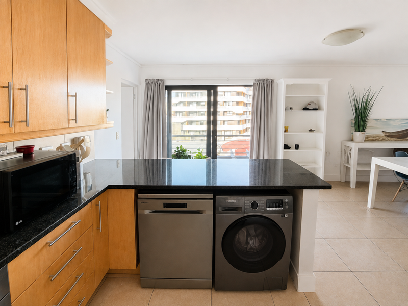 Modern kitchen featuring light wood cabinetry, dark granite countertops, and integrated laundry and dishwasher appliances, opening into a bright living area.