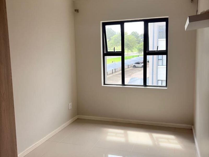A bright, empty bedroom featuring neutral tiled flooring, built-in cupboard storage, and a large window with black aluminium frames.