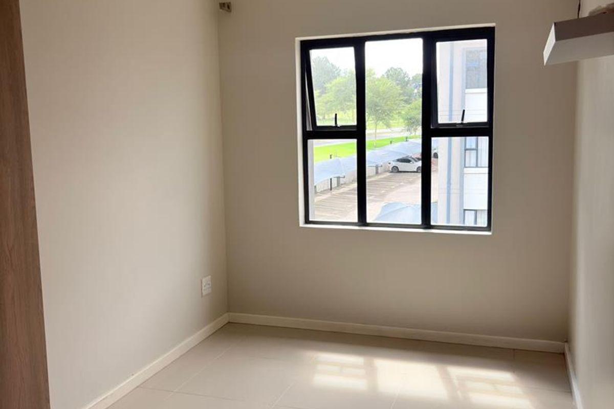 A bright, empty bedroom featuring neutral tiled flooring, built-in cupboard storage, and a large window with black aluminium frames.