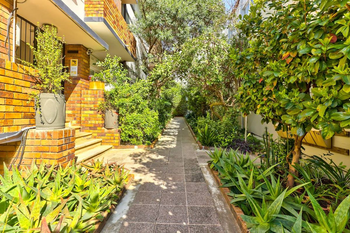 A well-maintained exterior walkway featuring brick building facades, paved pathways, and established garden landscaping.