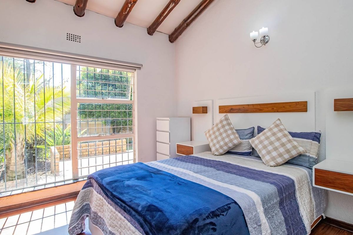 A bright bedroom featuring exposed timber ceiling beams, a custom built-in headboard with floating bedside tables, and large windows with security bars.