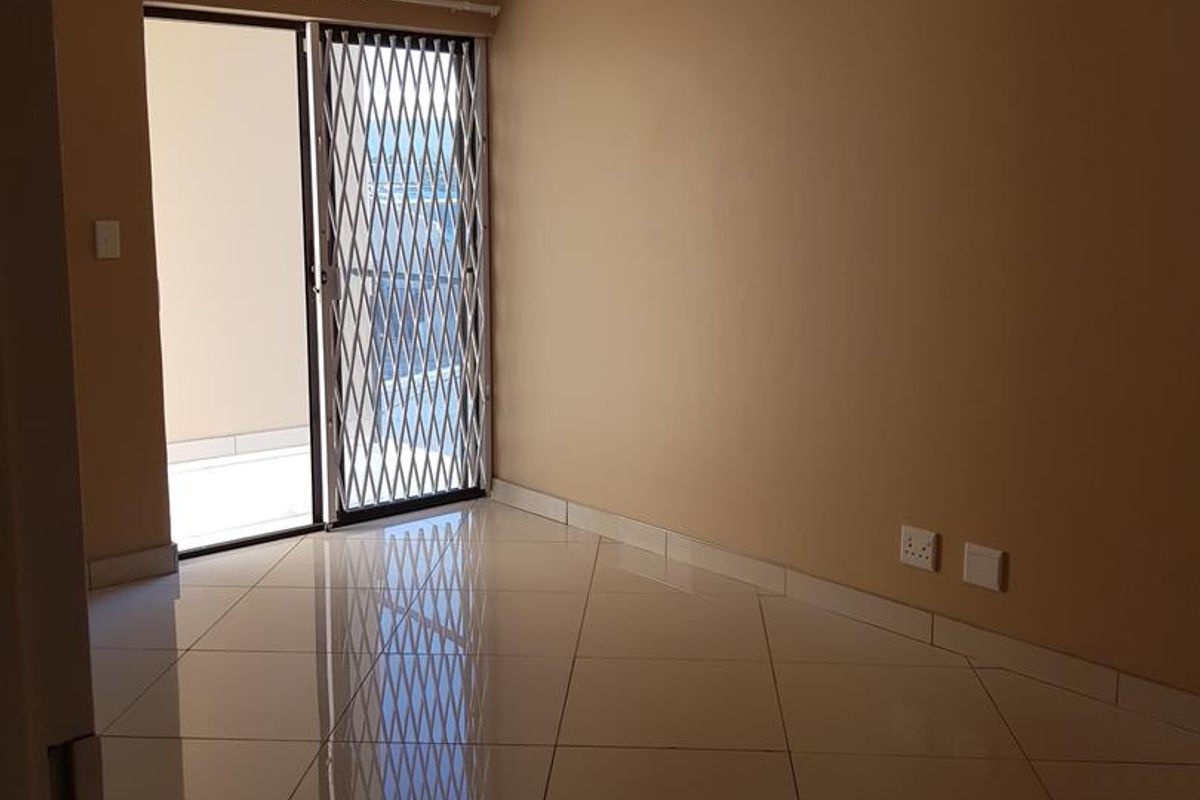 An empty living room featuring polished floor tiles, a ceiling light fixture, and a sliding glass door with a security gate leading to a balcony.