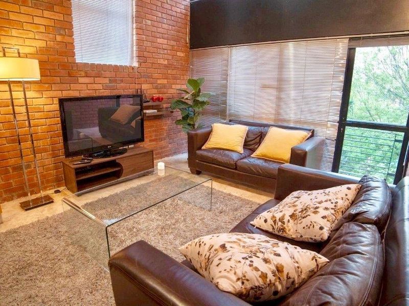 A living room featuring an exposed brick feature wall, large windows with venetian blinds, and access to an outdoor balcony area.