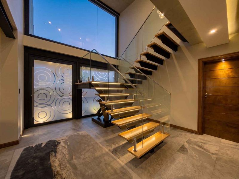 Modern entrance hall featuring a striking floating staircase with glass balustrades, integrated LED lighting, and large format floor tiles under a double-volume ceiling.