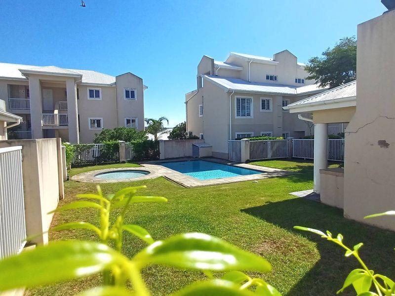 Communal outdoor area featuring a swimming pool, spa, and lawn, surrounded by apartment building facades and perimeter fencing.