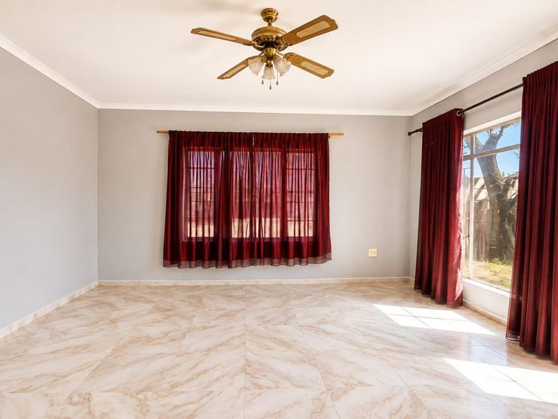 A bright, empty living room featuring neutral tiled flooring, a ceiling fan, and large windows that provide ample natural light.