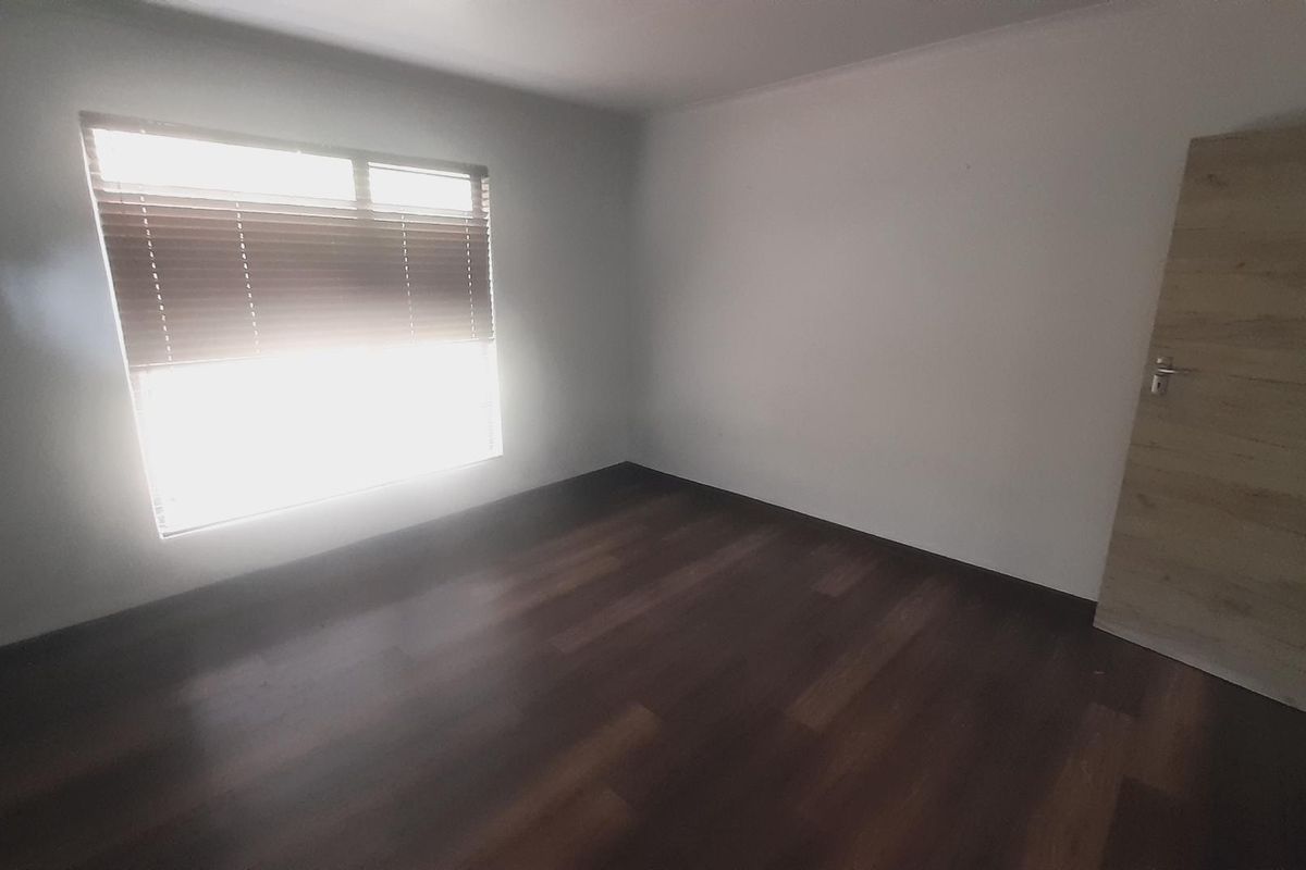 A bright, empty bedroom featuring dark wood-look flooring, neutral walls, and a window fitted with horizontal blinds.
