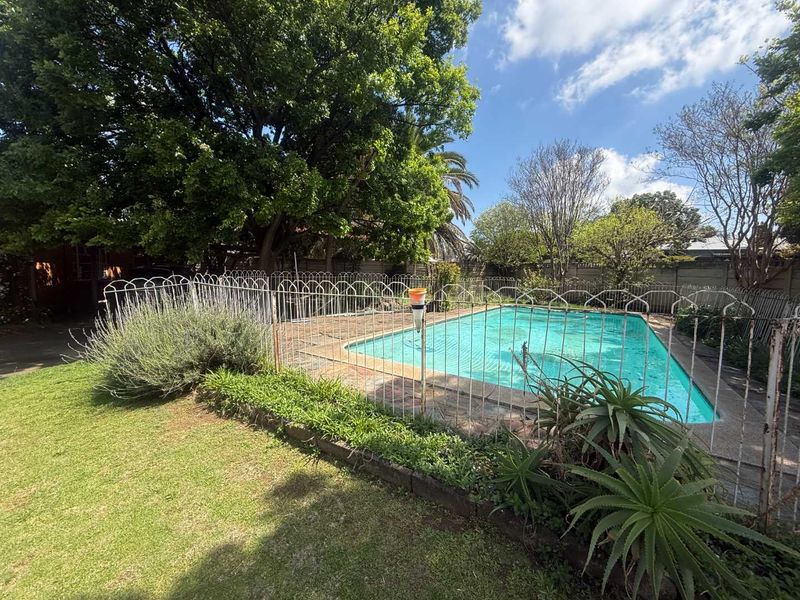 Outdoor pool area featuring a swimming pool enclosed by metal fencing, set within a mature garden with established trees and lawn.