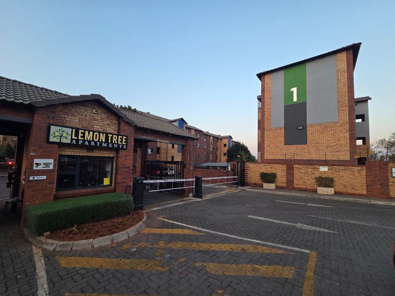 The entrance to Lemon Tree Apartments featuring a secure boom gate, brick facade architecture, and paved driveway access.