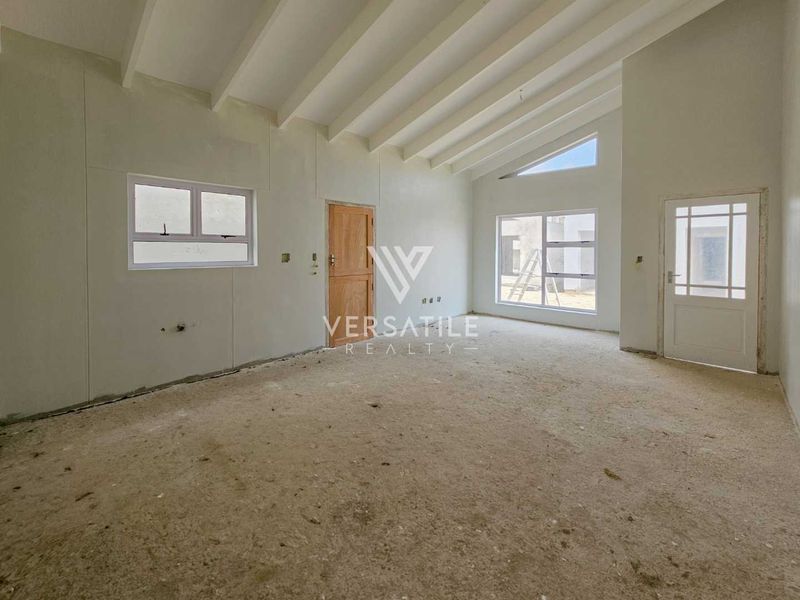 An unfinished, open-plan living area featuring high exposed beam ceilings, large windows, and neutral wall finishes, awaiting final flooring and interior detailing.