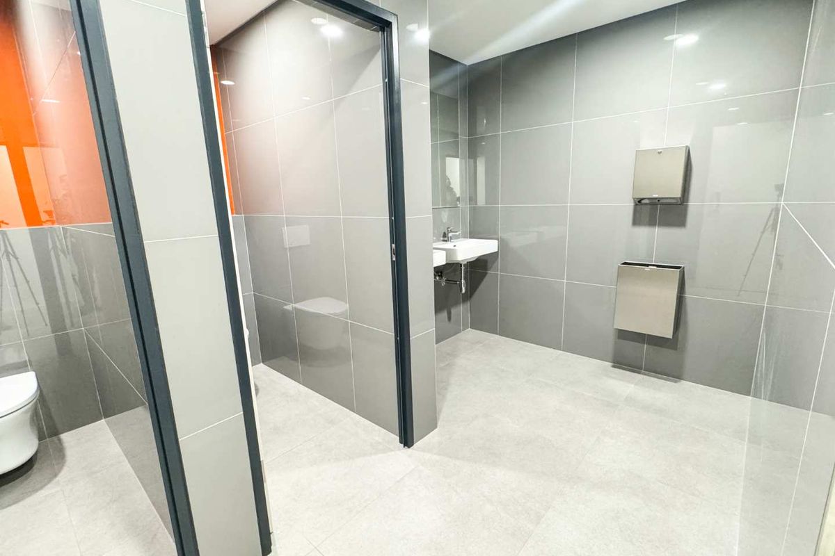A modern, clean commercial bathroom featuring grey tiled walls and floors, a wall-mounted sink, and stainless steel fixtures.
