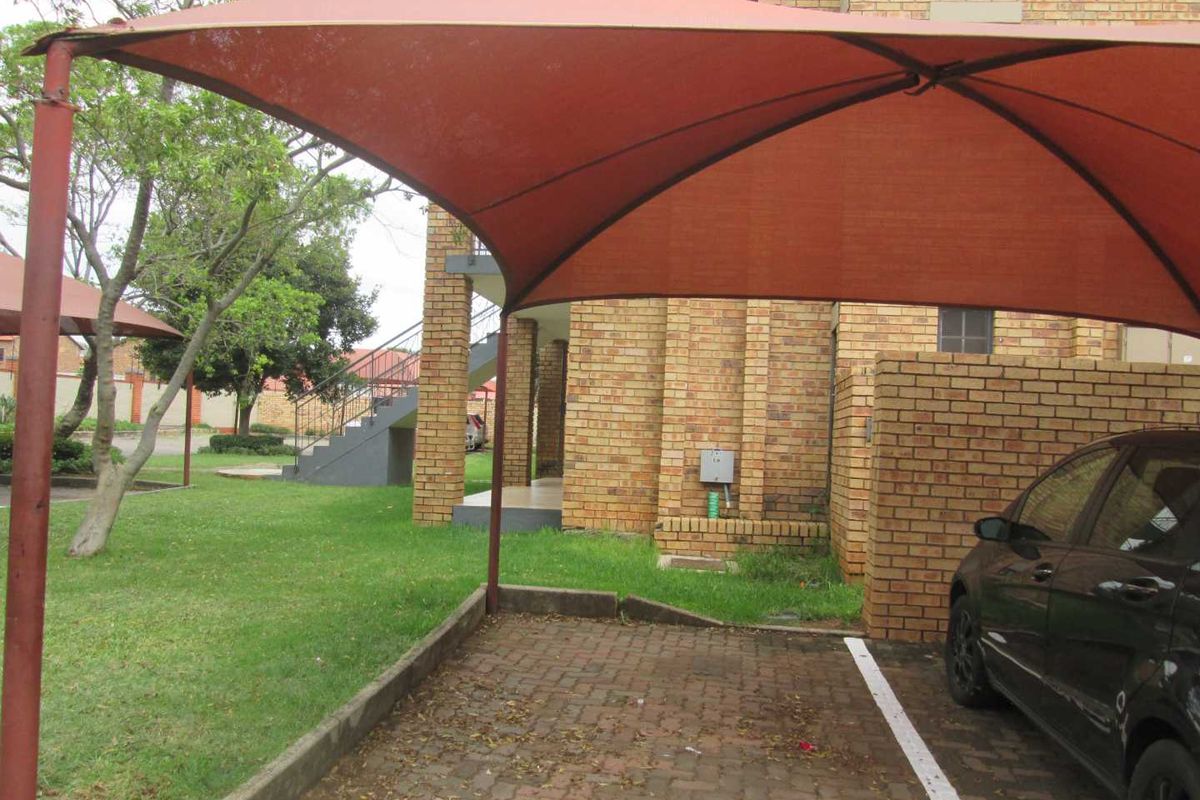 Covered parking bay with brick paving and shade cloth structure, situated within a residential apartment complex with landscaped garden areas.