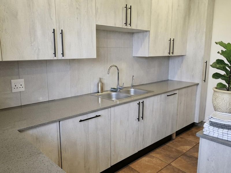 A functional scullery featuring light wood-grain cabinetry, stone countertops, a double bowl sink, and tiled finishes.