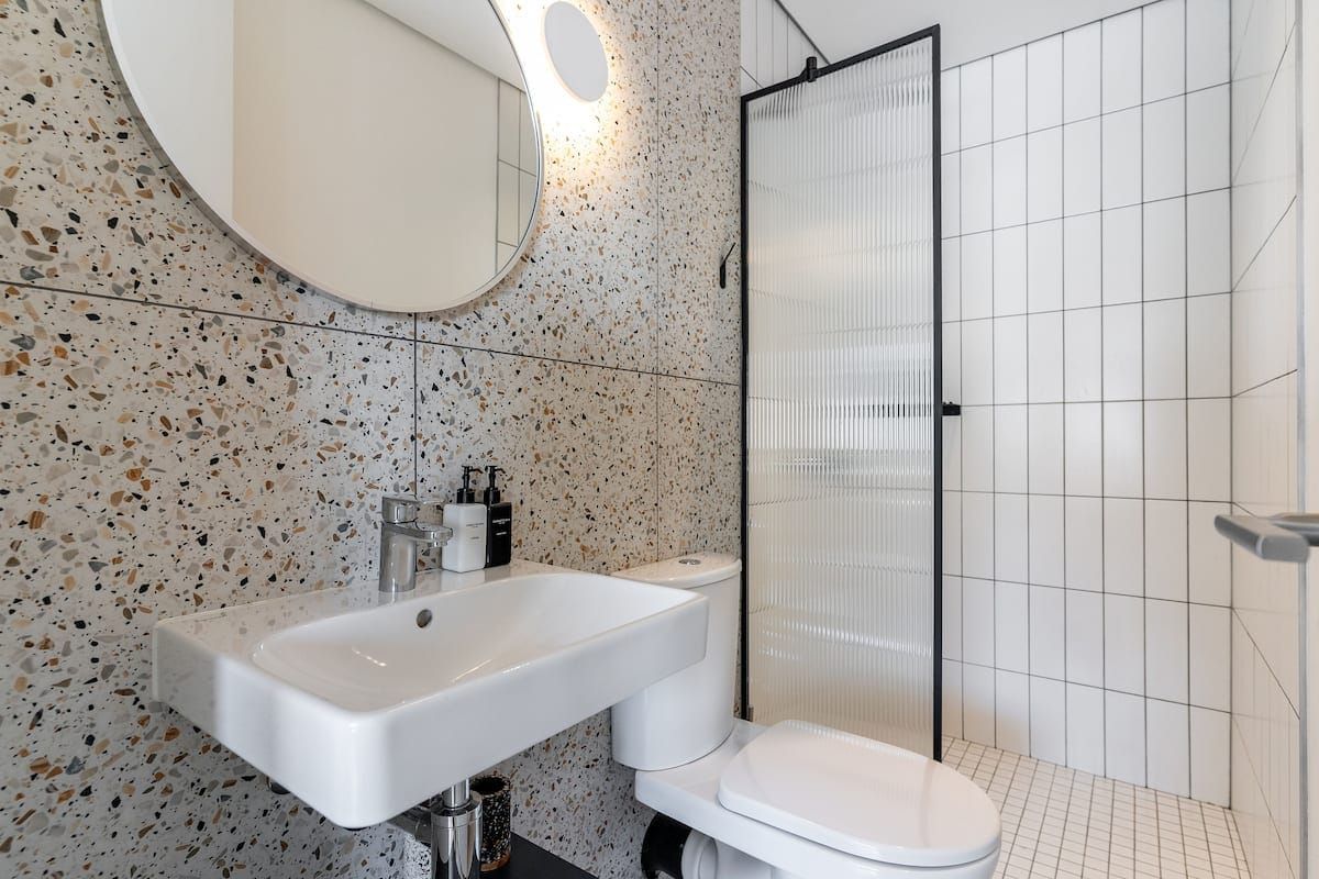 Modern bathroom featuring terrazzo-style wall tiles, a wall-mounted basin, and a walk-in shower with a fluted glass screen and black frame.