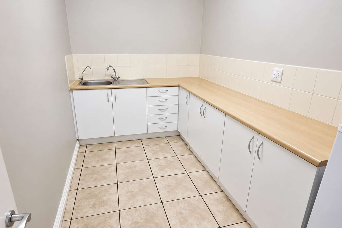 A clean and functional kitchenette featuring white cabinetry, light wood-look laminate countertops, a stainless steel sink, and neutral ceramic floor tiling.