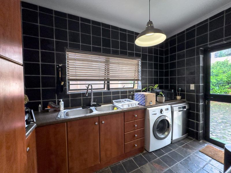 Functional kitchen featuring dark tiled walls and floors, wooden cabinetry, double sink, and integrated plumbing for laundry and dishwasher appliances.