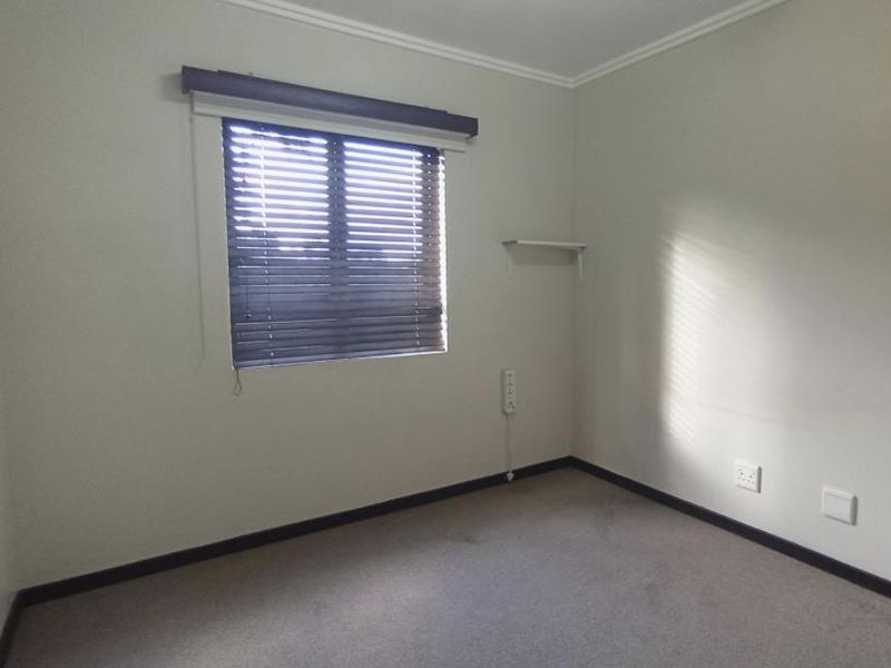 A neutral, empty bedroom featuring carpeted flooring, a window with venetian blinds, and wall-mounted electrical points.